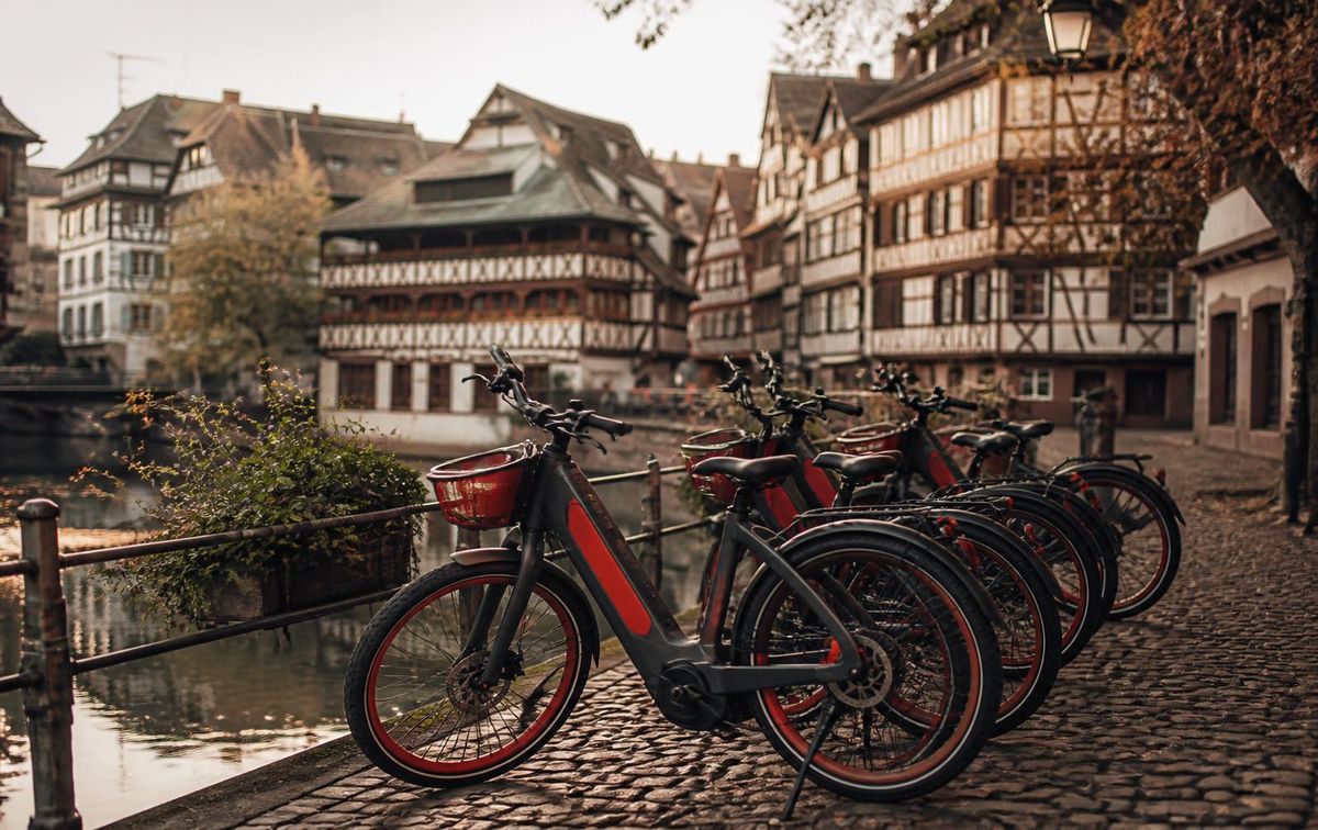 Eine Reihe rot akzentuierter E-Citybikes auf einem Kopfsteinpflaster-Kai in der Petite France in Straßburg, dahinter Fachwerkhäuser