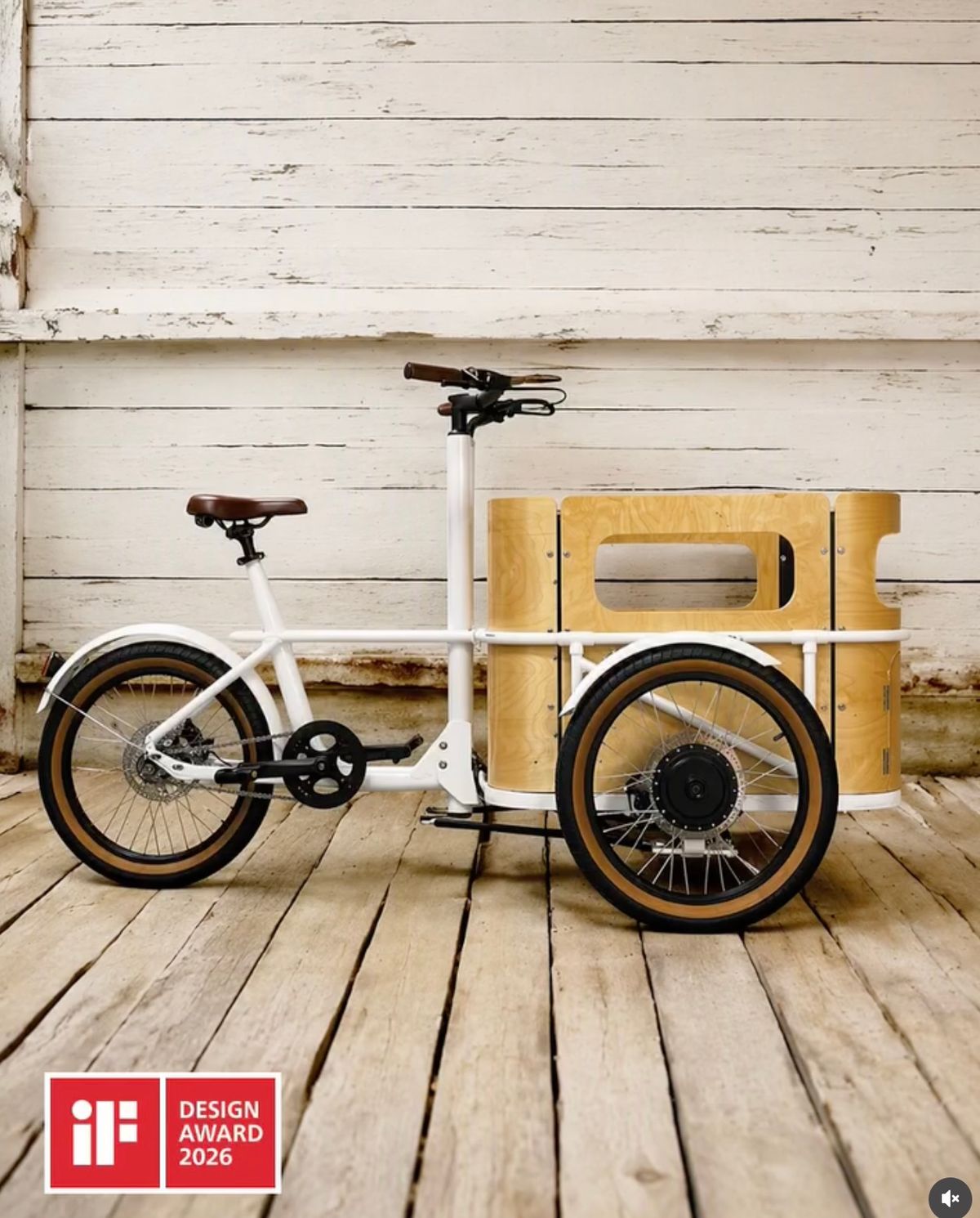 Das weiße minica E-Lastenrad mit Birkensperrholz-Box, ausgezeichnet mit dem iF Design Award 2026, vor einer verwitterten weißen Holzwand fotografiert