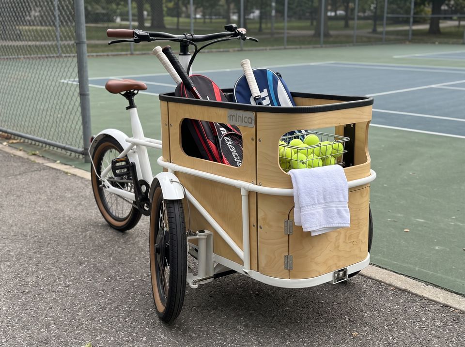 Ein minica E-Lastendreirad mit Schlägertaschen und einem Korb Tennisbälle am Platz.